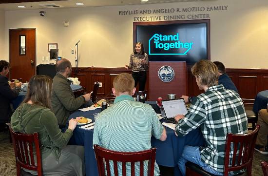 students at roundtables with stand together logo behind guest speaker